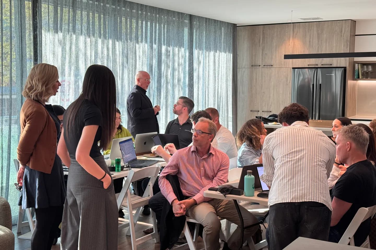 A workshop in a client's office: people at laptops around trestle tables, two facilitators standing at the front, floor-to-ceiling windows behind sheer curtains.