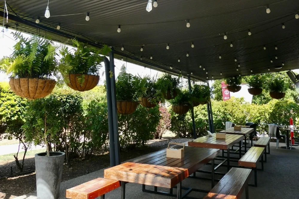 The Mayfair Emerald's outdoor dining area: timber tables and benches under a shade roof strung with festoon lights, hanging ferns along the edge.