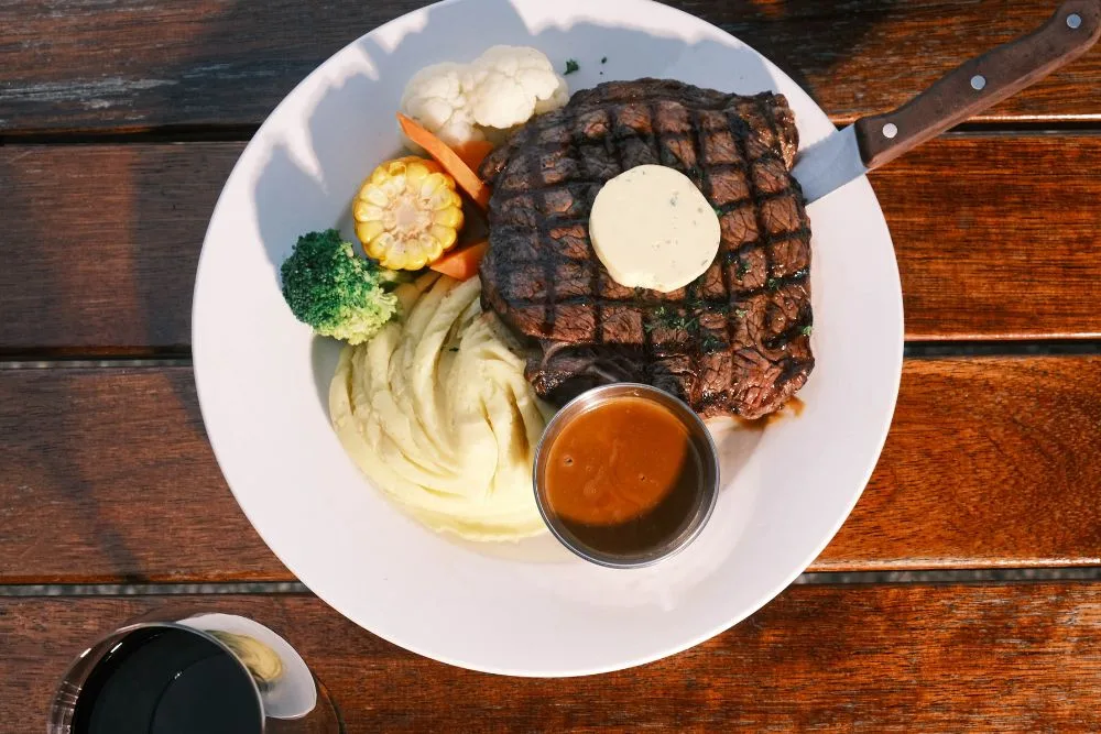 A plate from The Mayfair Emerald's bistro: a grilled steak with herb butter, mashed potato, corn, broccoli and cauliflower, a pot of gravy, and a glass of red wine on the timber table.
